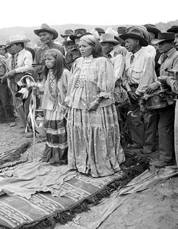 A group of Indigenous people, including two girls in patterned dresses, stand together outdoors on woven blankets, surrounded by men in hats.