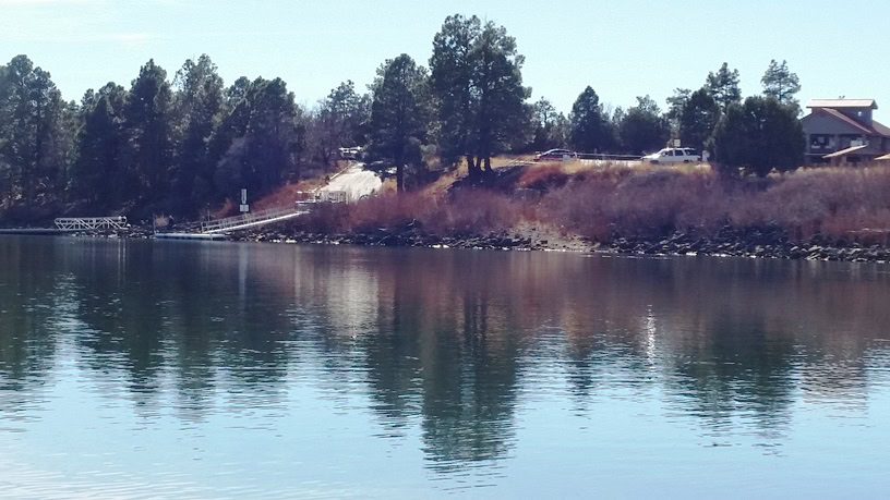 A calm lake reflects trees and a house on a hill; a dock extends into the water near the shoreline under a clear sky.