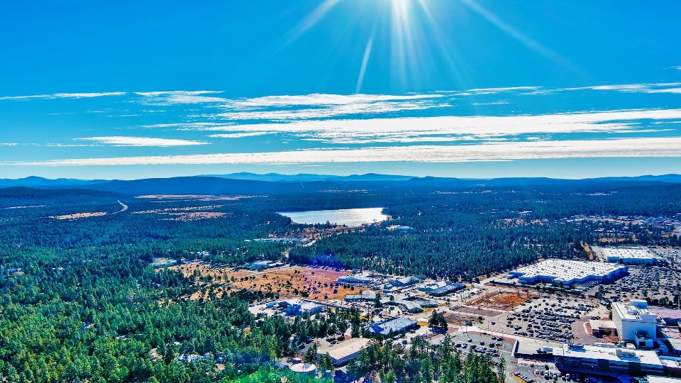 Aerial view of a small town surrounded by forested areas, a large lake, distant mountains, and a clear blue sky with the sun shining brightly.