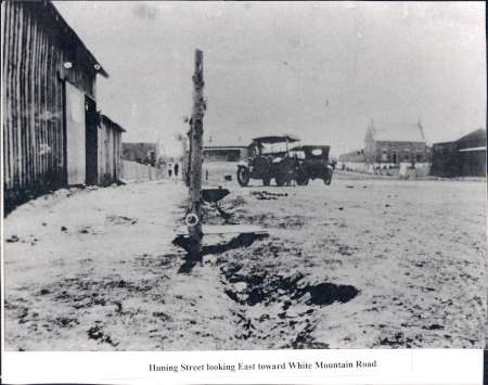 Black-and-white photo of an unpaved street with a wooden pole in the foreground, old cars, and buildings on both sides. Sign at bottom reads: "Huning Street looking East toward White Mountain Road.