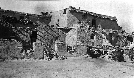 Black and white photo of a multi-story adobe building with stone and mud walls, featuring visible stairs, doorways, and exposed wooden beams.