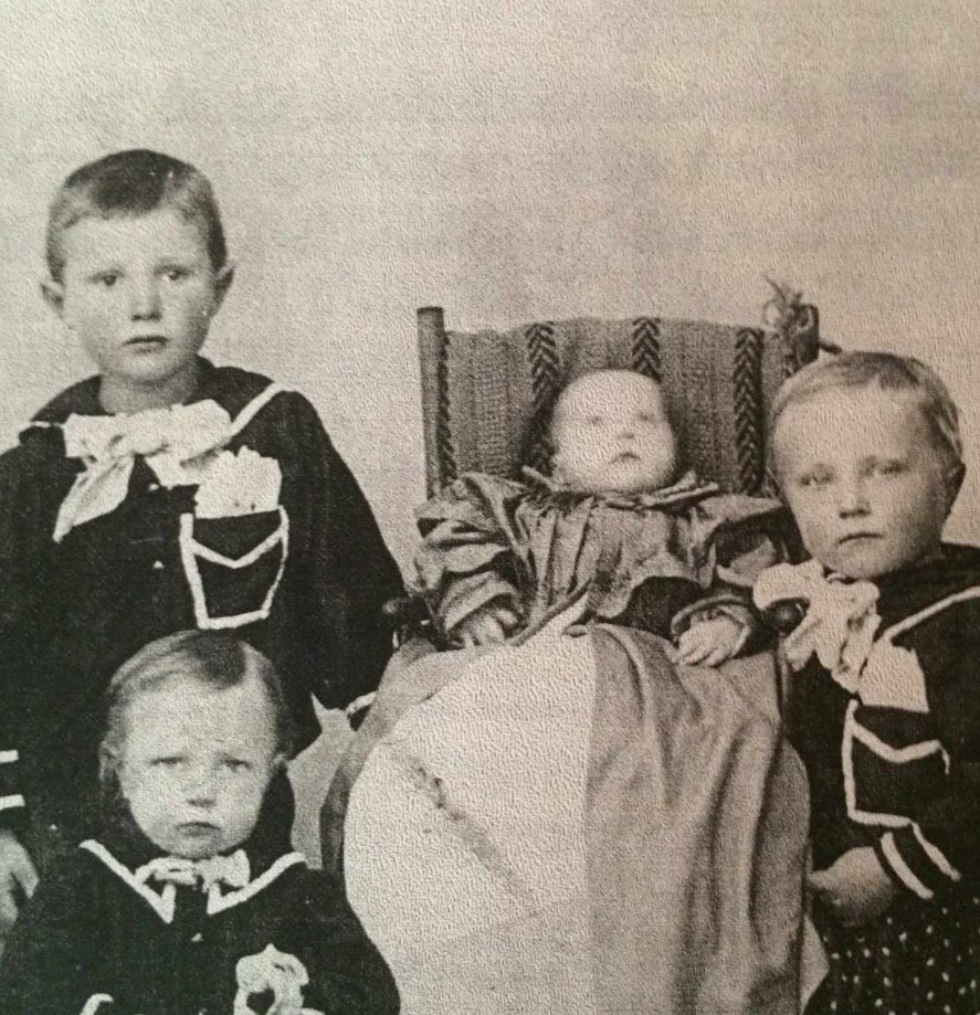 Four young children, three sitting and one in a chair, dressed in old-fashioned clothing, pose for a black-and-white portrait.