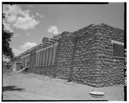Single-story stone building with wooden beams and boarded windows, set on a dirt ground under a partly cloudy sky.