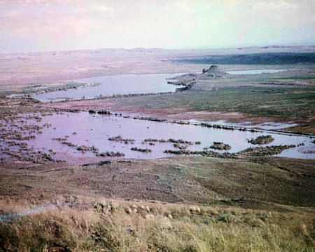A view of a partially flooded landscape with patches of water, grassy terrain, and distant hills under a cloudy sky.