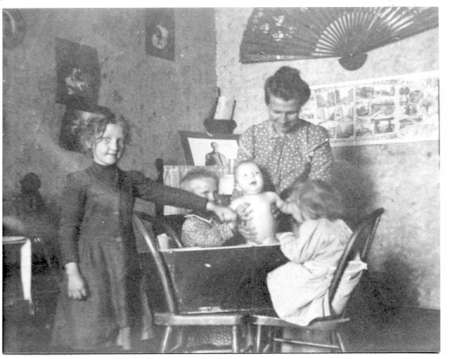 A woman stands behind three children and a baby seated around a table in a room decorated with a wall fan and pictures.