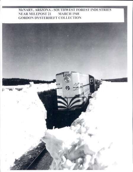 A train travels through deep snow on cleared tracks near McNary, Arizona, in March 1948; snowbanks rise high on either side of the train.