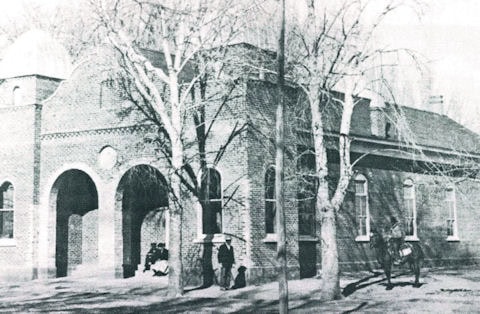 Black and white photo of a brick building with arched entryways and bare trees in front; several people stand or sit near the entrance.