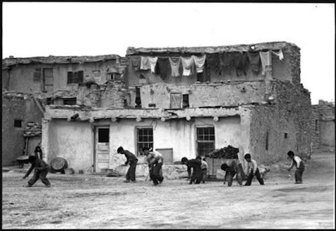 Children play a game in front of an old two-story adobe building with laundry hanging on the upper level.