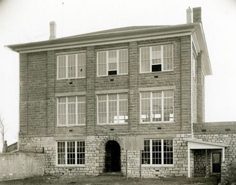Black-and-white photo of a three-story stone building with large windows, arched doorway at the center, and a small attached structure on the right side.