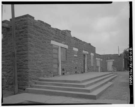 Black and white photo of a stone building with missing roof and several doorways, fronted by wide steps leading up from the street.