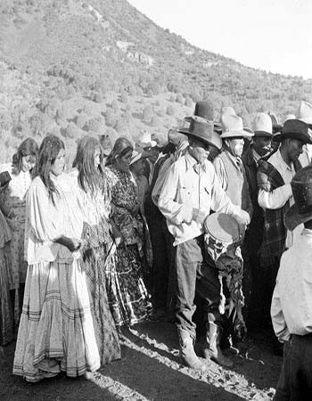 A group of Indigenous people, some wearing hats and traditional clothing, stand outdoors in a line near a mountain, with a man playing a drum in the foreground.