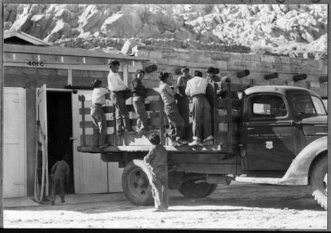 A group of children stand and sit on the back of a flatbed truck parked beside a building with an open door; rocky terrain is visible in the background.