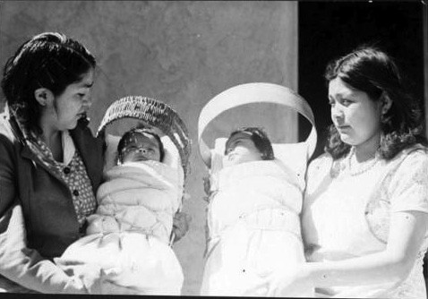 Two women each hold a bundled baby in a carrier, standing side by side and looking at the infants.