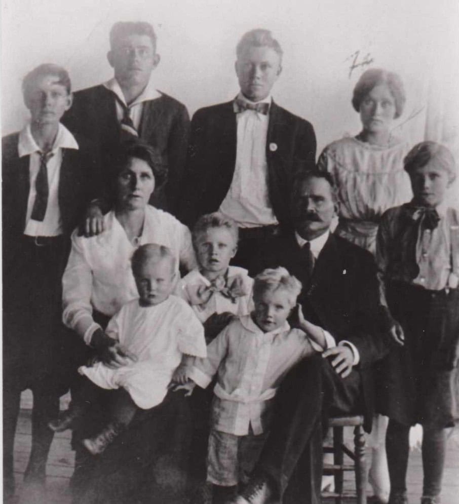 Black and white photo of a family with seven children and two adults, posed indoors in formal attire, with some seated and others standing.