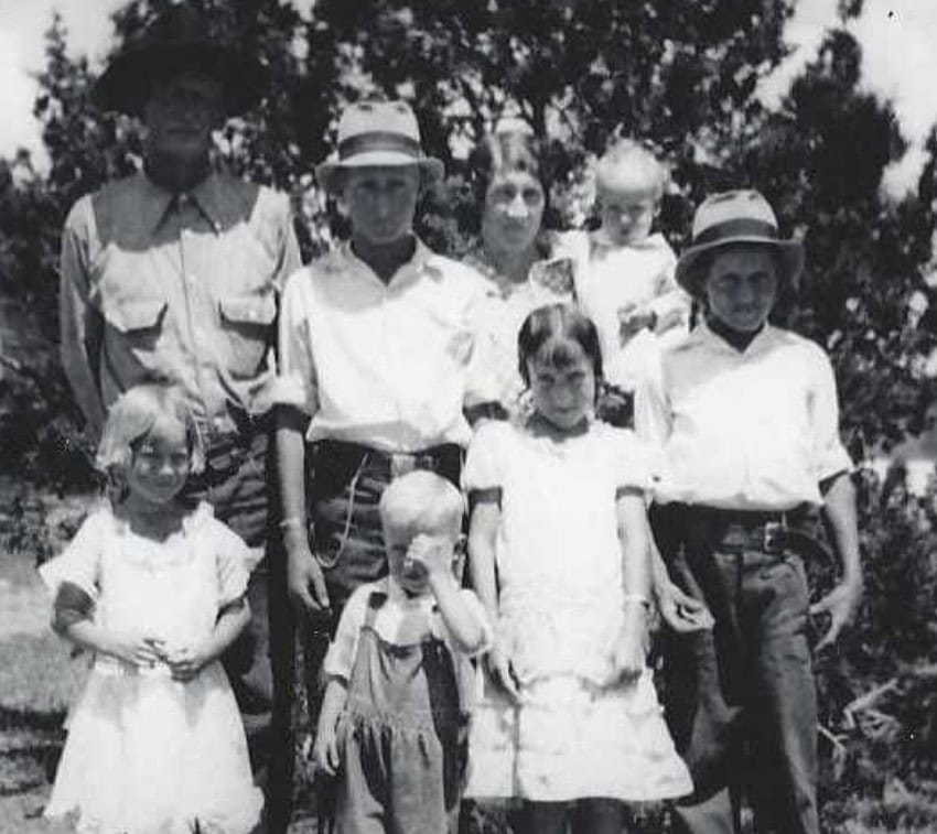 Black and white photo of a family of eight, including two adults and six children, standing outdoors in front of trees.