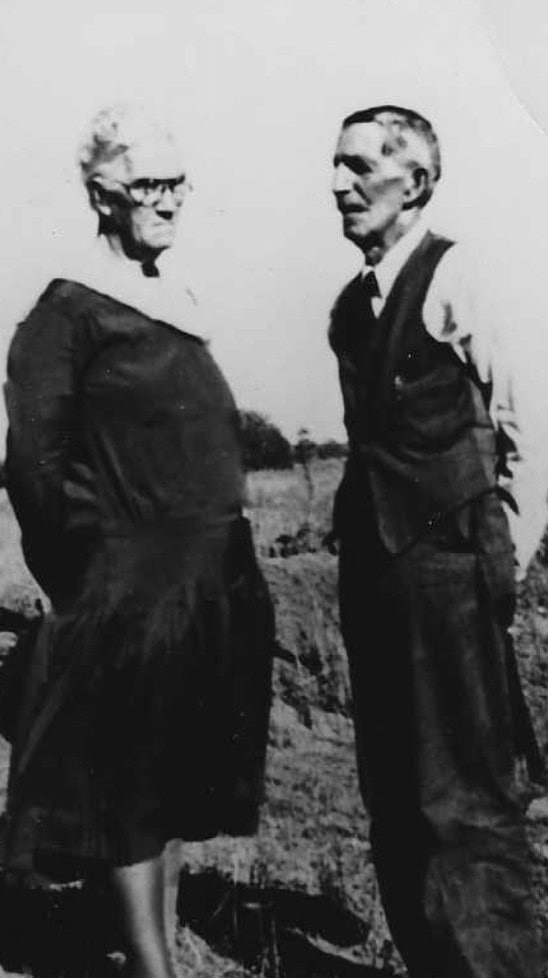An older woman and man stand outdoors in a grassy area, facing each other. The photo is in black and white.