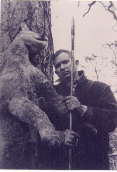 A man stands next to a large, upright bear with a spear in hand, outdoors near a tree.