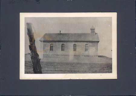 Black-and-white photo of a simple rectangular building with three arched windows and a small cupola, set in a barren landscape with a tree in the foreground.