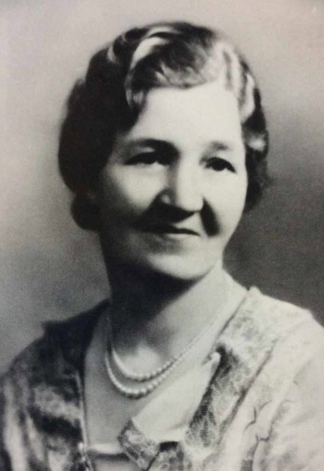 Black and white portrait of a woman with short, wavy hair, wearing a light-colored blouse and a pearl necklace, facing slightly to the right and smiling gently.