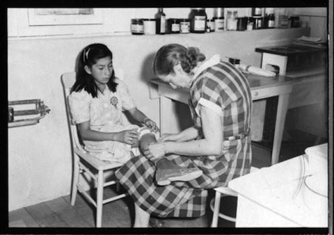 A woman in a plaid dress bandages a young girl's leg in a medical clinic setting with shelves of bottles in the background.