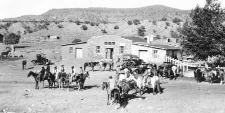 Historic black-and-white photo of people on horseback and early cars gathered outside a stone building in a rural, hilly landscape.