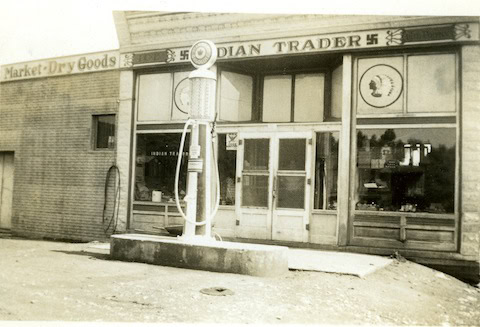 Black-and-white photo of an old storefront labeled "Indian Trader" with a vintage gas pump in front; swastika symbols are visible on the building sign.