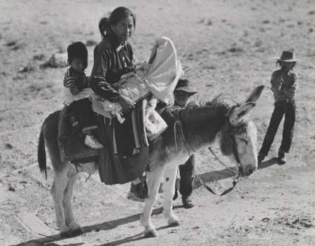 A woman carrying two children sits on a donkey in a dry, rural landscape, with two men standing nearby.