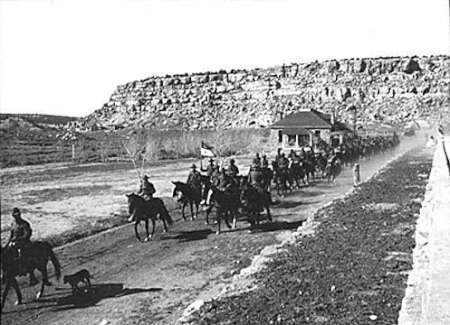 A group of mounted soldiers ride along a road near a rocky hillside, passing a small building, with dust rising behind them.