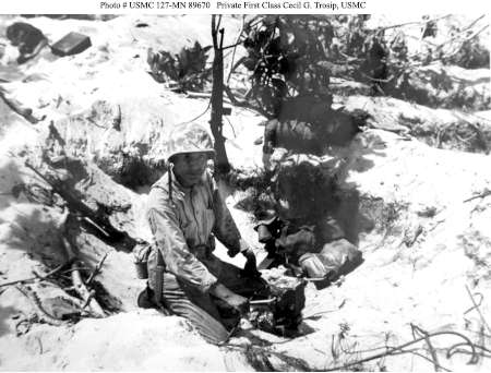 A soldier in uniform sits in a shallow trench or foxhole on sandy terrain with some equipment and vegetation around him.