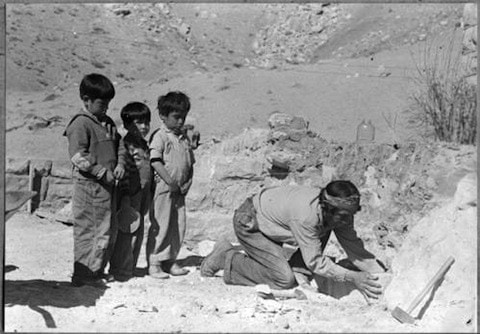 Four young boys stand in a line, watching an adult kneeling and working with tools on the ground in a rocky, outdoor setting.