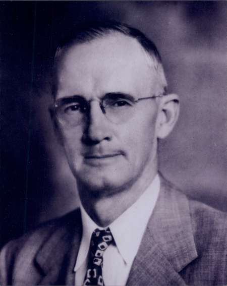 Black and white portrait of an older man wearing glasses, a suit, and a patterned tie, facing the camera with a neutral expression.