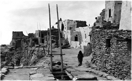 Black and white photo of a person walking along a path beside adobe and stone buildings in a cliffside village.