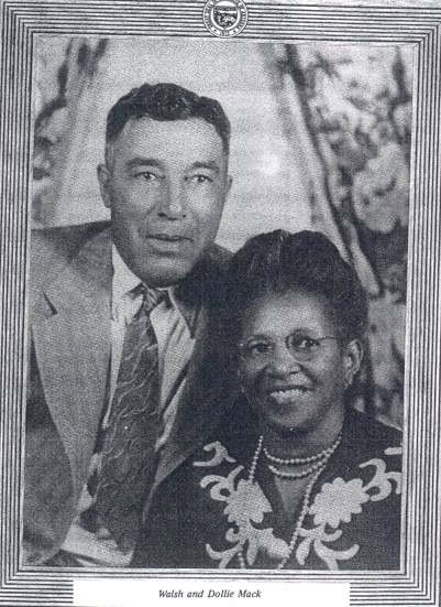 Black-and-white portrait of a man in a suit and a woman in glasses and a dress with floral patterns, seated side by side and smiling. The caption below reads "Walsh and Dollie Mack.