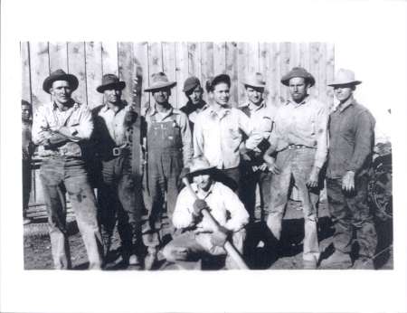 Black and white photo of nine men standing and one man kneeling in front of a wooden wall, all wearing work clothes and hats.