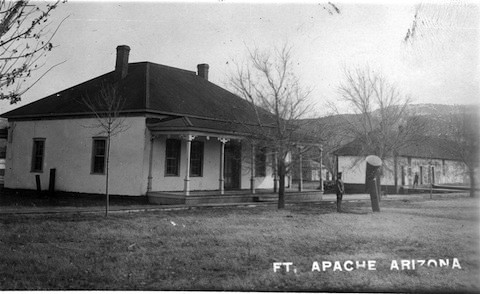 Black and white photo of two buildings at Fort Apache, Arizona, with barren trees and two people standing in front.