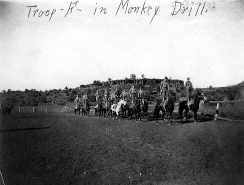 A group of uniformed soldiers on horseback ride in formation outside on a dirt field, with "Troop H in Monkey Drill" written on the photo.