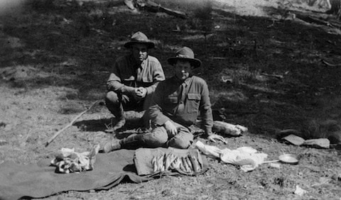Two men in outdoor clothing sit on the ground with caught fish and supplies laid out on a blanket in front of them, in a forested area.