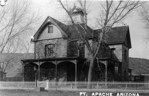 Historic stone building with a covered porch and cupola, surrounded by a white picket fence and bare trees, labeled "FT. APACHE ARIZONA.