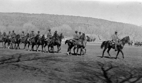 A group of soldiers on horseback rides in formation across an open field with hills and trees in the background.