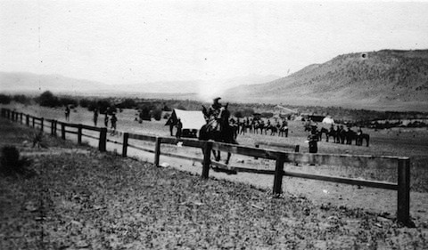 Black and white photo of a person on horseback near a wooden fence, with tents and a group of people or horses in the background on open land.