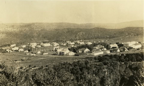 A black-and-white photo shows a small town or settlement with scattered buildings surrounded by open fields and distant hills.