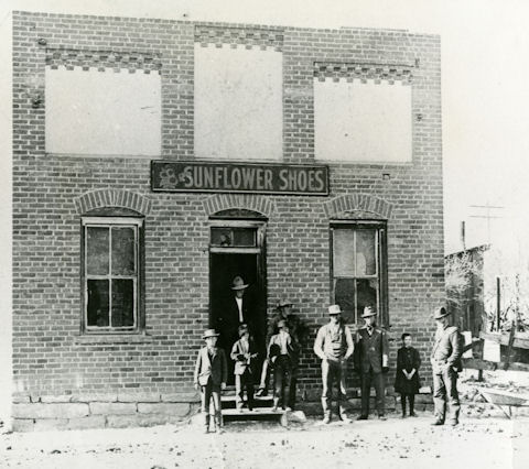 Black-and-white photo of people standing in front of a brick building with a sign reading “Sunflower Shoes.” The group includes men, boys, and a girl.