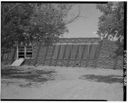 Black-and-white photo of a stone building with several large windows, shaded by trees, and a ramp leading to one window.
