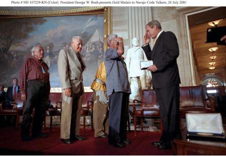 President George W. Bush presents a medal to a Navajo Code Talker, who salutes him, with others waiting in line in a large hall with statues and a mural, July 26, 2001.
