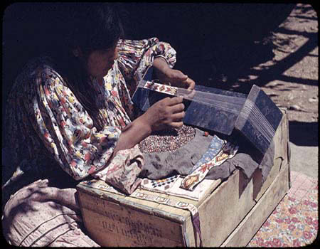 A person sits outdoors, using a loom to weave a colorful patterned textile on top of a wooden box.