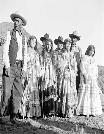 A group of Indigenous men and women, some in traditional dress, stand outdoors in a row with a mountain in the background.