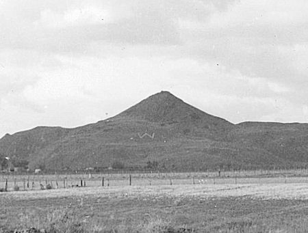 A hill with a large white "W" symbol on its slope, surrounded by a flat field and scattered fences under a cloudy sky.
