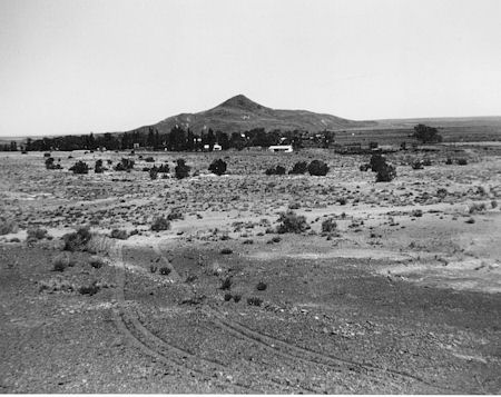 Black and white photo of a dry, sparsely vegetated landscape with a low, rounded hill in the background and a few small buildings and trees clustered at its base.