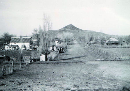 Black and white photo of a rural dirt road lined with fences, small houses on either side, and a hill in the background.