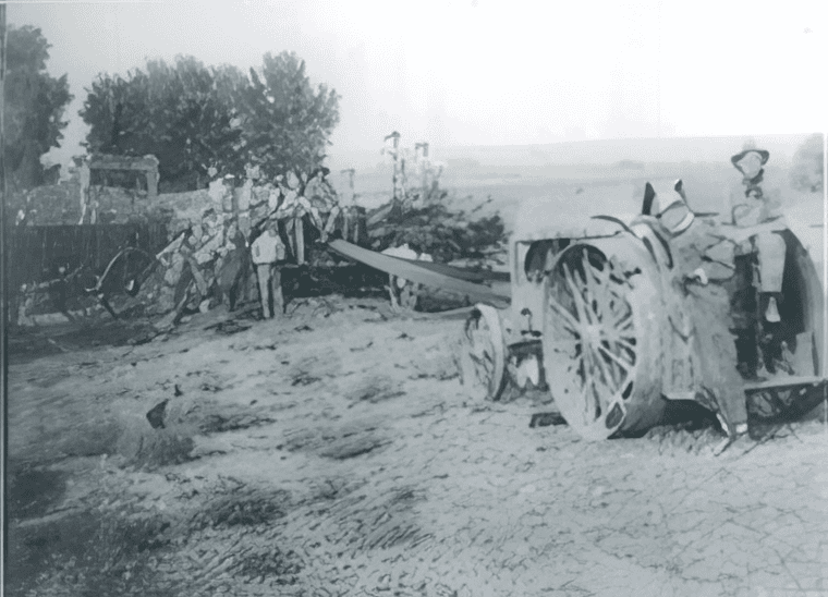 Black and white photo of several people operating a large farming machine attached to a tractor in a rural landscape with trees in the background.